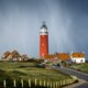 asphalt road between grass field going to brown lighthouse beside houses under cloudy sky