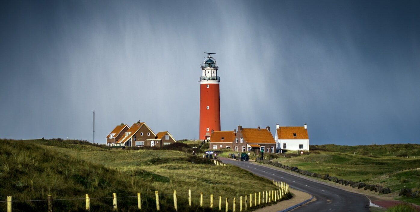 asphalt road between grass field going to brown lighthouse beside houses under cloudy sky